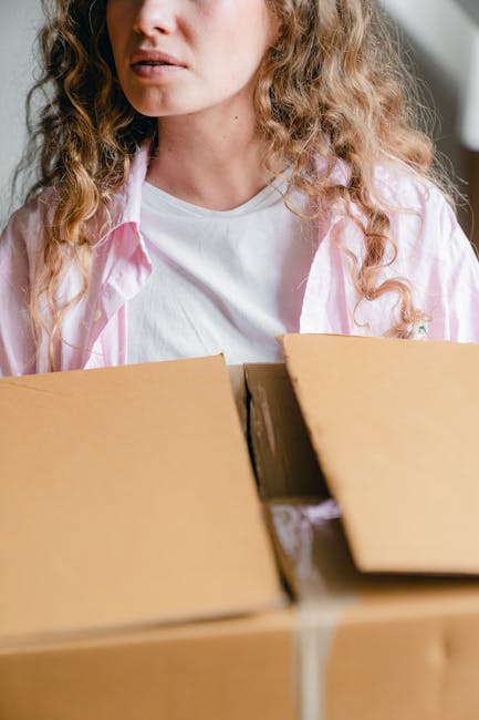 Close-up of a person in a white shirt holding a black clipboard with papers attached, standing next to stacked cardboard boxes and packing materials in an indoor setting. The individual appears to be 