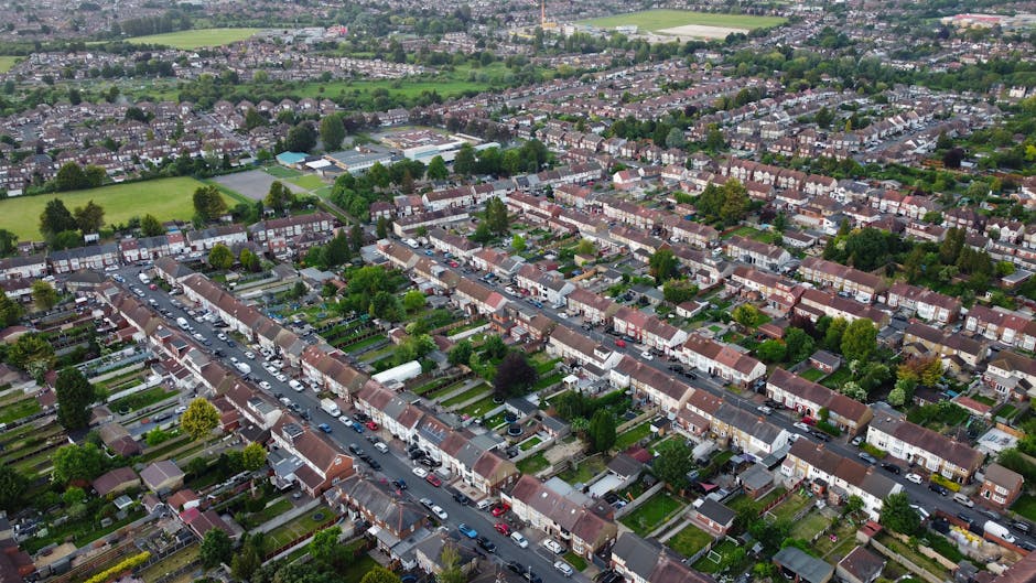 Aerial view of a residential neighbourhood in Canons Park, Edgware, showing rows of terraced houses with small gardens at the front and back, parked cars lining the streets, and lush green trees scattered throughout the area. In the background, there are sports fields and open green spaces, with a mix of detached and semi-detached houses extending into the distance. The scene captures a typical suburban setting, with clear pathways and roads in a well-organised layout, exemplifying a peaceful community environment suitable for house removals and home relocation services offered by Edgware Removals, which specialize in furniture transport, packing, and moving logistics.