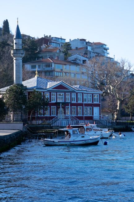 A close-up view of a small white motorboat with a cabin, docked near the edge of a body of water during daytime. The boat is secured with ropes to the dock and has red and white safety equipment visible on top. In the background, there is a red and white wooden house with large windows and a staircase leading to the entrance; the house is situated along a waterfront promenade with some trees nearby. Behind the house, a hilltop area features multi-story residential buildings with balconies, windows, and varied roof styles, under a clear blue sky. The scene captures a peaceful moment during a house removal or furniture transport process as part of a relocation service provided by Edgware Removals, showcasing typical elements of home relocation logistics near Edgware Station, HA8.