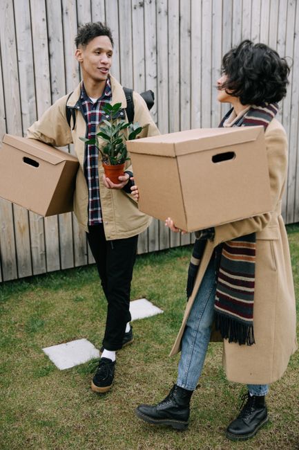 A young woman with curly, shoulder-length hair and fair skin, wearing a light blue denim shirt, stands inside a room filled with moving boxes of various sizes, mostly made of cardboard. Behind her, th