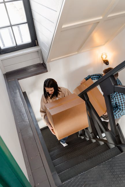 Interior view of a staircase inside a residential property during a home relocation, with two individuals carrying large cardboard moving boxes up the steps. One woman with dark hair and a beige top i